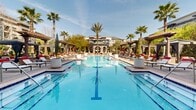 This two-floor property showcases an impressive resort-inspired outdoor amenity space as its centerpiece. The grand entrance features a covered porte-cochère with a decorative brick paver roundabout, flanked by vibrant tropical plantings and glass double doors set within a stone-clad facade — establishing a welcoming, upscale arrival experience.

The outdoor pool area is the defining highlight of the property. A large, sparkling lap-style pool with blue tile detailing anchors the space, surrounded by an expansive paved deck lined with cushioned lounge chairs accented in bold red. Towering palm trees are strategically placed throughout, providing natural shade and a tropical atmosphere. Multiple dark-canopied pergolas and private cabanas offer shaded retreats along the perimeter, creating intimate lounging areas within the broader communal space.

The layout of the outdoor area is thoughtfully designed with clear sightlines from the pool deck back to the building's facade, which features arched architectural detailing and balconies on the upper level. The brick and stone exterior of the surrounding structures adds warmth and visual texture to the environment. A secondary, more intimate pool or spa area is also present, surrounded by its own paved gathering space with additional seating and shade structures. Overall, the property exudes a vibrant, resort-caliber atmosphere ideal for outdoor relaxation and social gatherings.