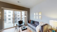 Located at 8900 Lyra Dr in Columbus, Ohio, this single-floor residence offers a thoughtfully designed living space with a clean, modern aesthetic. The living area serves as the heart of the home, showcasing hardwood-style flooring that adds warmth and continuity throughout the space. Neutral white walls are complemented by a bold accent wall in a deep taupe tone, creating a stylish visual contrast that adds depth to the room. Large sliding glass doors flood the interior with abundant natural light while providing direct access to a private balcony, offering views of the surrounding community. The open layout comfortably accommodates a generous seating arrangement, making it well-suited for both relaxing and entertaining. Overhead ventilation and modern lighting fixtures contribute to the functional design of the space. The overall ambiance is contemporary and welcoming, with a carefully curated palette of cool neutrals and muted tones that create a calm and sophisticated atmosphere. The combination of indoor comfort and outdoor connectivity makes this a versatile and appealing living space in the Columbus area.