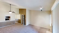 This two-story space offers a practical and straightforward layout designed for comfortable everyday living. The interior is characterized by a neutral color palette, with warm beige and earthy gold accent walls creating subtle visual interest throughout. Soft, wall-to-wall carpeting runs across the main living area, adding warmth underfoot and contributing to a cozy atmosphere. The textured ceiling adds a classic touch, while track lighting provides focused illumination to complement the overall brightness of the space. A half-wall partition with a pass-through opening adds an architectural element that defines separate areas while maintaining an open, connected feel between spaces. Doorways lead to additional rooms, hinting at a thoughtfully segmented floor plan. The staircase, partially visible, connects the two floors seamlessly, reinforcing the multi-level nature of the layout. Clean white baseboards and trim provide a crisp contrast against the carpeted floors and painted walls. Overall, the space presents a clean, move-in ready environment with a simple yet functional design, well-suited for a variety of uses and easily adaptable to a range of interior styles and preferences.