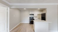 This thoughtfully designed one-bedroom, one-bathroom single-floor unit makes excellent use of its compact footprint. Light wood-style flooring runs throughout the entire space, creating a cohesive and clean aesthetic accented by neutral warm-gray walls and crisp white trim and crown molding.

The open living area flows directly into the kitchen, which features white upper and lower cabinetry, light stone-look countertops, a stainless sink, and black appliances including an electric range, range hood, and full-size refrigerator. A peninsula counter provides a natural boundary between the kitchen and living space. The living area is illuminated by recessed lighting and includes a window-unit air conditioner for climate control.

The bedroom is a comfortable, well-proportioned room featuring a ceiling fan with light fixture, a window with blinds that allows natural light, and direct access to both a walk-in closet and the bathroom. The walk-in closet is outfitted with multiple built-in shelving units. The bathroom includes a white vanity with a marble-look countertop, a framed mirror, and a shower-tub combination with white tile surround. This unit offers a practical and move-in-ready layout well-suited for single occupants or couples.