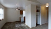 This single-floor space offers a functional and straightforward layout centered around an open-concept design that connects the kitchen and living areas. A partial wall divides the two zones, providing a sense of separation while maintaining an open, airy feel throughout.

The kitchen is equipped with warm-toned wood cabinetry that extends to the upper walls for ample storage, a white dishwasher, a white refrigerator, and a sink positioned beneath a window that allows natural light to filter in. The countertops offer practical workspace, and the tile flooring in the kitchen area adds durability and easy maintenance.

The adjacent living area transitions to carpet, creating a softer, more comfortable zone for relaxing. A ceiling fan with an integrated light fixture is centrally mounted overhead, providing both air circulation and ambient lighting throughout the shared space.

The walls are finished in a neutral, light tone that keeps the interior feeling bright and clean. The layout is practical and efficient, making good use of the available space. Overall, the space presents a simple, well-organized environment with functional finishes suited for everyday living.