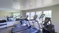 Located at 251 Eisenhower Dr in Biloxi, Mississippi, this fitness center offers a well-equipped, single-floor workout facility designed to support an active lifestyle. The space is outfitted with a lineup of modern cardio machines, including treadmills, ellipticals, and a stationary bike, all from reputable brands such as True Fitness. A full-length mirror runs along one wall, visually expanding the room and allowing users to monitor their form during exercise. Large, multi-pane windows line the opposite wall, flooding the space with abundant natural light and offering views of the outdoor surroundings. The clean, neutral-toned walls and recessed lighting contribute to a bright and welcoming atmosphere. A workout reference chart is posted on the wall, providing guidance for users of all fitness levels. The smooth, light-colored ceiling with recessed fixtures adds a polished, finished look to the room. The layout is thoughtfully organized to allow comfortable movement between equipment. Overall, this fitness center delivers a functional and inviting environment well-suited for daily cardio and general exercise routines.