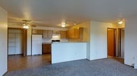 This single-floor space features an open-concept design that connects the kitchen and living area in a seamless, functional layout. The kitchen is equipped with warm wood-toned cabinetry providing ample upper and lower storage, a range with an overhead ventilation hood, and a full-size white refrigerator. A peninsula counter divides the kitchen from the living area, creating a natural transition between the two spaces while offering additional prep or casual dining surface. The kitchen floor is finished in tile, providing a durable, easy-to-clean surface, while the living area features wall-to-wall carpet that adds warmth underfoot. Track lighting runs along the kitchen ceiling, supplemented by a flush-mount ceiling fixture, ensuring the space is well-lit throughout. The walls are painted in a neutral tone, contributing to a bright and clean atmosphere. The open flow between the kitchen and living area makes the space feel connected and practical, well-suited for everyday living. Overall, the layout prioritizes functionality while maintaining a cohesive and comfortable ambiance throughout.