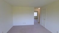 This single-floor space features a straightforward, functional layout with neutral design elements throughout. The main room is finished with wall-to-wall carpeting in a light taupe tone, creating a soft, understated foundation. Crisp white walls provide a clean, blank canvas adaptable to a variety of furnishing styles. A doorway on the right side of the room leads to an adjacent space, while an open passage connects to a secondary room visible in the background, which benefits from a window allowing natural light to filter through. A flush-mount ceiling light fixture in the connecting hallway area provides overhead illumination. Baseboard heating is present along the walls of the adjoining room, indicating a practical heating setup throughout. Electrical outlets are positioned along the walls at standard height. The overall design is simple and unadorned, with consistent finishes carried throughout the visible areas, creating a cohesive and neutral atmosphere. The space is presented vacant, allowing for a clear view of its structural layout and flow between rooms.