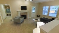 This single-floor space offers a welcoming and functional layout centered around a comfortable living area. The room is anchored by a classic white-mantled fireplace with tile surround, above which a wall-mounted flat-screen television is positioned for convenient viewing. Crown molding lines the upper edges of the warm, neutral-toned walls, adding a touch of traditional architectural detail throughout the space.

Natural light fills the room through multiple windows fitted with horizontal blinds, creating a bright and airy atmosphere. A ceiling fan overhead provides both illumination and air circulation, contributing to the room's everyday practicality. The living area is furnished with contemporary seating, including a pair of light gray armchairs and a tufted dark sofa, arranged around a round accent table — a setup that encourages relaxed conversation and leisure.

The flooring transitions between carpet in the seating area and hard surface tile, offering both comfort and durability. A glass-paneled exterior door provides additional natural light and access to the outdoors. The open layout connects smoothly to an adjacent area, enhancing the overall sense of space and flow throughout this well-appointed, single-level property.