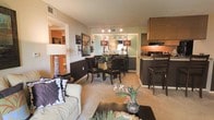 This single-floor residence offers a thoughtfully designed open-concept layout that seamlessly connects the living, dining, and kitchen areas into one cohesive and functional space. The living area is anchored by a plush sofa accented with decorative pillows, complemented by a sleek coffee table and warm, neutral-toned carpeting that runs throughout. Adjacent to the living space, the dining area features a glass-top table surrounded by dark wood chairs, with a mirrored accent wall and stylish lamp fixtures adding a touch of elegance. The kitchen is positioned conveniently nearby, showcasing dark cabinetry, granite-style countertops, and a raised breakfast bar with seating — ideal for casual dining or entertaining. Track lighting in the dining nook highlights the space beautifully, while the warm color palette of teal, brown, and tan tones creates a cohesive and inviting ambiance throughout. A hallway visible from the main living area suggests additional private rooms beyond, completing this well-proportioned and stylishly appointed single-level residence.
