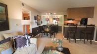 This single-floor residence offers a thoughtfully designed open-concept layout that seamlessly connects the living, dining, and kitchen areas into one cohesive and functional space. The living area is anchored by a plush sofa accented with decorative pillows, complemented by a sleek coffee table and warm, neutral-toned carpeting that runs throughout. Adjacent to the living space, the dining area features a glass-top table surrounded by dark wood chairs, with a mirrored accent wall and stylish lamp fixtures adding a touch of elegance. The kitchen is positioned conveniently nearby, showcasing dark cabinetry, granite-style countertops, and a raised breakfast bar with seating — ideal for casual dining or entertaining. Track lighting in the dining nook highlights the space beautifully, while the warm color palette of teal, brown, and tan tones creates a cohesive and inviting ambiance throughout. A hallway visible from the main living area suggests additional private rooms beyond, completing this well-proportioned and stylishly appointed single-level residence.