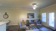 This single-story space presents a well-appointed living area that combines comfort with contemporary style. The room features elegant crown molding along the ceiling perimeter, adding a refined architectural touch, while a ceiling fan with a built-in light fixture provides both function and subtle charm overhead.

The living area is anchored by a plush gray sofa accented with patterned throw pillows, complemented by a matching armchair that adds additional seating. A set of industrial-style nesting coffee tables in wood and metal sits atop a geometric area rug, grounding the space with a modern aesthetic. Dark hardwood-style flooring runs throughout, providing a rich contrast against the neutral-toned walls.

Natural light filters in through a window on the right side of the room, brightening the space during daytime hours. A decorative tripod floor lamp adds a warm, ambient glow, while a potted plant in the corner introduces a touch of greenery. Wall art depicting an urban streetscape serves as a tasteful focal point, contributing to the overall modern and inviting character of the living space.
