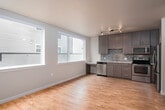 This single-floor residence offers a thoughtfully designed layout that balances functionality with modern style. The open living area is bright and airy, featuring light hardwood flooring and large windows that allow natural light to fill the space while offering views of the surrounding neighborhood. A bold gray accent wall adds a contemporary touch and visually defines the living zone, complemented by clean white trim and neutral wall tones throughout.

The kitchen is a standout feature, outfitted with a full suite of stainless steel appliances including a range, over-the-range microwave, refrigerator, and dishwasher. Sleek flat-front cabinetry in a warm wood grain finish pairs elegantly with granite countertops and a mosaic tile backsplash in muted blue and gray tones. A built-in desk area adjacent to the kitchen provides a convenient workspace bathed in natural light from a large window.

The bathroom continues the refined aesthetic with a dark wood vanity, granite countertop, undermount sink, and a well-lit vanity mirror above. Light hardwood flooring extends into the bathroom, maintaining a cohesive feel throughout the unit. The overall design emphasizes clean lines, warm materials, and a neutral palette, creating a comfortable and modern living environment.