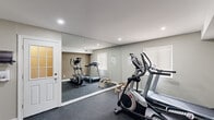 Located at 310 Shipley Rd in Wilmington, Delaware, this dedicated home fitness room is a thoughtfully designed single-level space that maximizes every square foot for an efficient workout environment. The room is finished with durable dark rubber flooring, ideal for high-impact exercise and equipment placement, while neutral taupe-toned walls create a clean, focused atmosphere throughout.

A standout feature is the floor-to-ceiling mirror wall that spans a significant portion of the room, visually expanding the space and providing the functional benefit of form-checking during workouts. Recessed ceiling lighting ensures bright, even illumination across the entire area. A window fitted with blinds introduces natural light while maintaining privacy.

The space is well-appointed with a range of cardio equipment, including an elliptical trainer, a stationary exercise bike, and a treadmill. A dedicated alcove along one wall serves as an organized storage area, housing a tiered dumbbell rack with varying weights, a stability ball, and additional fitness accessories. A flat workout bench positioned in the open floor area adds versatility for strength training exercises.

A security camera is mounted near the ceiling, and the room includes an interior door for convenient access. The overall layout is practical and functional, making it a comprehensive private gym suitable for a full range of cardiovascular and strength training workouts.
