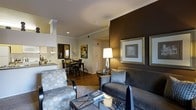 This single-floor residence features an open-concept layout that seamlessly connects the living area, dining space, and kitchen. The living room is anchored by a bold dark brown accent wall that adds depth and contrast to the otherwise warm, neutral color palette throughout the space. Crown molding runs along the ceiling, lending a refined architectural detail to the interior. The room is furnished with a plush charcoal sofa accented by decorative pillows, complemented by a patterned accent chair and a glass-top coffee table. A sleek table lamp provides warm ambient lighting, while framed wall art enhances the sophisticated aesthetic. Rich hardwood-style flooring flows throughout, adding warmth and continuity between spaces. The dining area, visible in the background, accommodates a dark wood table and chairs, ideal for everyday dining. Adjacent to the dining area, the kitchen features light cabinetry and appears well-equipped for daily use. Recessed lighting and a neutral wall palette keep the space feeling bright and cohesive. Overall, the interior presents a contemporary, well-appointed environment with a balanced mix of comfort and style.