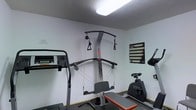 This dedicated fitness space offers a practical and functional environment for personal exercise, all contained on a single floor. The room features light-colored walls and a textured ceiling with bright overhead fluorescent lighting, creating a clean and well-lit atmosphere conducive to focused training sessions.

The centerpiece of the space is a wall-mounted MAX cable pulley system, complete with dual adjustable cables, a bench, and a variety of attachments — enabling a wide range of strength and resistance exercises. A treadmill is positioned nearby, providing a convenient option for cardiovascular workouts, while a stationary exercise bike rounds out the cardio equipment offerings.

A workout reference chart is posted on the wall, serving as a practical guide for exercise routines. The flooring appears to be a dark, durable material suited for gym use. The overall layout is efficiently arranged to maximize the usable area while keeping all essential equipment accessible.

This space is well-suited for full-body training, combining strength and cardio equipment in a straightforward, no-frills setting designed to support consistent and effective workouts.