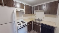 This single-floor unit offers a straightforward, functional layout with distinct living, sleeping, kitchen, and bathroom areas. The kitchen is equipped with dark wood-toned cabinetry spanning both upper and lower storage, light-colored countertops, white tile backsplash, a coil electric range, refrigerator, dishwasher, and a single-basin sink. Wood-look vinyl flooring runs through the kitchen area, adding a practical, easy-to-maintain surface.

The living area is generously proportioned, featuring neutral-toned carpet and a sliding glass door fitted with vertical blinds that allows natural light to fill the space and provides access to an outdoor area. A baseboard heating system lines the perimeter walls throughout the unit. The bedroom is a well-sized, carpeted room with a window dressed with horizontal blinds, offering natural light and baseboard heating. The bathroom features a full-size soaking tub with a fully tiled white surround, maintaining a clean and classic appearance. Overall, the unit presents a practical and livable layout with functional finishes throughout.