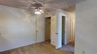 This single-floor residential space is designed with practical functionality in mind. The bedroom is a standout feature, finished with plush wall-to-wall carpeting that adds warmth and comfort underfoot. A ceiling fan with an integrated light fixture is centrally mounted on the textured ceiling, providing both airflow and ambient lighting throughout the room. The room benefits from generous built-in storage solutions, including a dedicated closet with a hanging rod for clothing and an adjacent open shelving unit that offers versatile organizational options for a variety of items. A secondary door visible in the background suggests easy connectivity to additional areas of the home, contributing to a practical and flowing layout. The neutral color palette of white walls and beige carpeting creates a clean, understated aesthetic that is easy to personalize. The textured ceiling adds a subtle design element consistent throughout the space. Overall, the room presents a straightforward and well-equipped living environment, with storage and comfort as central priorities, making it a functional and adaptable space for everyday residential use.