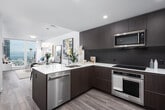 This contemporary single-floor residence offers a thoughtfully designed open-concept layout that seamlessly connects the kitchen, dining, and living areas. The kitchen is outfitted with dark wood cabinetry, stainless steel appliances including a built-in oven, microwave, and refrigerator, complemented by a striking marble-look quartz countertop island with an undermount sink and warm gold hardware. Light wood flooring flows throughout, enhancing the bright, airy atmosphere. The open living and dining area is bathed in natural light from floor-to-ceiling windows, with the dining space anchored by a round table and the living area featuring a neutral-toned sofa alongside bold artwork. The bedroom is a serene retreat, with a slatted wood accent wall, a bold navy feature wall, and a large window framing city skyline views, flooding the room with natural light. A well-organized walk-in closet with built-in shelving and hanging rails provides generous storage. The bathroom is clean and minimalist, showcasing a frameless glass walk-in shower with white subway tile surround and polished chrome fixtures. Throughout the residence, the design palette of whites, warm woods, and muted blues creates a cohesive, modern aesthetic.