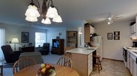 This single-floor residence offers a thoughtfully designed open-concept layout that seamlessly connects the main living areas. The kitchen is outfitted with light wood cabinetry, ample storage, and a central island with a white countertop that doubles as a breakfast bar, complete with bar stool seating. A white refrigerator and additional upper cabinets provide generous storage, while tile flooring anchors the space with a clean, practical finish. Adjacent to the kitchen, the dining area features a wooden table and chairs beneath an elegant multi-light chandelier with frosted glass shades, adding a classic decorative touch to the space. The living area flows naturally from the dining space, featuring plush carpeting, a cozy recliner, and a traditional wood-framed entertainment unit. A bright window along the exterior wall allows natural light to filter into the room, creating a welcoming ambiance. A dedicated laundry area is conveniently accessible from the main living space, adding to the home's functional appeal. Throughout the residence, warm wood tones, neutral wall colors, and a cohesive design palette contribute to a comfortable and inviting atmosphere suited for everyday living.