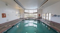 Located at 32905 19th Pl S in Federal Way, Washington, this single-floor indoor aquatic facility offers a well-appointed swimming and relaxation environment. The centerpiece of the space is a large indoor swimming pool with brick-tiled edging, complemented by an adjacent hot tub or spa. The pool area is bathed in natural light streaming through a sweeping curved bank of multi-pane windows that span a significant portion of one wall, reaching toward the ceiling and providing views of the surrounding greenery. The high ceilings contribute to a bright, open atmosphere throughout the space. The pool deck features durable textured flooring designed for wet environments, with safety rails providing convenient pool entry and exit points. A sliding glass door along one wall offers direct access to an outdoor area, enhancing ventilation and connectivity to the exterior. The facility also includes clearly marked safety equipment, including a pool shut-off switch, as well as informational signage and bulletin boards mounted along the walls. An interior window overlooking an adjacent room, decorated with colorful displays, suggests the facility is part of a larger community or multi-use building. The overall design prioritizes functionality, safety, and comfort for recreational aquatic use.