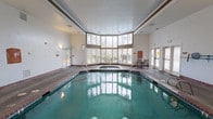 Located at 32905 19th Pl S in Federal Way, Washington, this single-floor indoor aquatic facility offers a well-appointed swimming and relaxation environment. The centerpiece of the space is a large indoor swimming pool with brick-tiled edging, complemented by an adjacent hot tub or spa. The pool area is bathed in natural light streaming through a sweeping curved bank of multi-pane windows that span a significant portion of one wall, reaching toward the ceiling and providing views of the surrounding greenery. The high ceilings contribute to a bright, open atmosphere throughout the space. The pool deck features durable textured flooring designed for wet environments, with safety rails providing convenient pool entry and exit points. A sliding glass door along one wall offers direct access to an outdoor area, enhancing ventilation and connectivity to the exterior. The facility also includes clearly marked safety equipment, including a pool shut-off switch, as well as informational signage and bulletin boards mounted along the walls. An interior window overlooking an adjacent room, decorated with colorful displays, suggests the facility is part of a larger community or multi-use building. The overall design prioritizes functionality, safety, and comfort for recreational aquatic use.