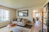 This single-floor residence offers a thoughtfully arranged layout that balances comfort and functionality throughout. The living room serves as the heart of the home, featuring rich hardwood-style flooring, a tiled fireplace with a wall-mounted television above, and large windows fitted with vertical blinds that invite abundant natural light and views of the surrounding greenery. A oversized framed mirror adorns one wall, enhancing the sense of space and brightness in the room. The open flow connects the living area to a dedicated dining space, creating an ideal setting for both relaxing and entertaining. The kitchen is equipped with warm wood cabinetry, granite-style countertops, a full suite of white appliances, and recessed lighting. A notable feature is the in-unit laundry area, conveniently integrated within the kitchen space and accessible via louvered doors. Two well-sized bedrooms offer plush carpeting, large windows with natural light, and tasteful wall art, providing comfortable and tranquil retreats. The bathroom is clean and functional, complete with a combination tub and shower, wood-style flooring, and a vanity with matching wood cabinetry. Throughout the residence, a neutral color palette creates a cohesive and inviting atmosphere.