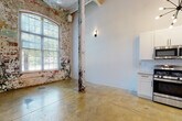 This single-floor industrial loft showcases a compelling fusion of historic building fabric and contemporary design. Throughout the space, polished concrete floors provide a durable, cohesive foundation, while soaring ceilings with exposed ductwork, original cast-iron columns, and weathered brick walls preserve the authentic character of the building's industrial past. Large arched windows along the exterior wall flood the main living areas with abundant natural light and offer views of the surrounding greenery.

The kitchen is thoughtfully appointed with white shaker cabinetry, subway tile backsplash, granite countertops, and a full suite of stainless steel appliances, including a gas range, microwave, dishwasher, and refrigerator. A striking starburst chandelier adds a stylish focal point overhead. The bathroom features a navy vanity with a granite countertop, undermount sink, matte black fixtures, and a large mirror. Additional rooms offer flexible open spaces defined by clean white walls that contrast with the raw structural elements, including original columns and exposed overhead piping. A long internal corridor, lined with wall sconces and housing the electrical panel and HVAC access, connects the various areas of the unit efficiently. The overall ambiance is an artful balance of rugged, reclaimed aesthetics and refined, move-in-ready amenities.