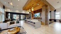 This well-appointed single-floor clubhouse blends warm natural materials with contemporary design to create an inviting, resort-style atmosphere. The central gathering area serves as the heart of the space, anchored by a lengthy white quartz bar counter lined with black-and-gold barstools. Behind it, a built-in wet bar with cabinetry, a sink, and a wall-mounted television is framed by richly textured wood paneling and a geometric tile backsplash. A wood-clad ceiling with pendant lighting in brushed gold adds warmth overhead.

Adjacent to the bar area, a comfortable lounge features tufted white sofas, velvet accent chairs, and an area rug, all bathed in natural light from floor-to-ceiling windows with botanical-print drapery. Tall ceilings and expansive glazing carry throughout the space, lending an open, airy quality.

A dedicated game room offers shuffleboard and billiards tables beneath a shiplap ceiling, while a separate private office provides a quiet workspace with double-height windows and plush seating. High-top bistro tables with gold pedestal bases occupy a bright transitional lounge area near large windows overlooking lush outdoor greenery. The entry foyer includes a self-serve coffee station set against decorative stonework and bold artwork, creating a welcoming arrival experience.