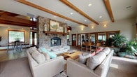 Located at 21580 East Bitterroot Lane in Liberty Lake, Washington, this single-floor community space is thoughtfully designed to blend rustic charm with modern functionality. The centerpiece of the main gathering area is a dramatic floor-to-ceiling stone fireplace, framed by exposed wooden ceiling beams that lend warmth and architectural character to the open, airy room. Recessed lighting throughout complements the natural light streaming in through large windows and glass-paned doors.

The open-concept layout connects the living lounge seamlessly to a dining area, where a generous dining table sits beside a window with views of lush greenery. The well-equipped kitchen features warm wood cabinetry, dark countertops, mosaic tile backsplash, stainless steel appliances, and tile flooring — designed for both practicality and style.

A dedicated fitness center rounds out the amenities, offering a comprehensive range of equipment including treadmills, ellipticals, cable machines, free weights, stability balls, and suspension training systems. A roll-up garage door opens the fitness space directly to the outdoors, providing fresh air and natural views during workouts.

Additional spaces include a casual game or media room with natural light and a relaxed atmosphere, as well as a welcoming lounge entry with leather seating beside the double-sided stone fireplace.