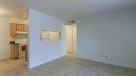 This single-floor space offers a clean, straightforward layout designed for functional living. The main living area is finished with light-toned tile flooring that extends throughout, lending a cohesive and easy-to-maintain surface underfoot. The walls are painted crisp white, contributing to a bright and airy atmosphere throughout the unit.

A notable architectural detail is the rectangular pass-through window cut into the dividing wall between the living area and the kitchen. This opening allows natural sightlines into the kitchen, where a white refrigerator is visible, and helps maintain a sense of openness between the two spaces without fully removing the wall separation.

Overhead, HVAC vents are integrated into the ceiling, ensuring climate control across the space. A hallway visible from the main living area leads toward additional rooms, with a classic louvered bi-fold closet door providing storage access. The overall ambiance is minimal and neutral, offering a blank canvas that can be adapted to a variety of interior styles. The space is well-suited for those seeking a straightforward, low-maintenance living environment.