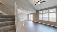 This two-story home is designed with an open, airy feel that is immediately apparent upon entering the main living area. The living room commands attention with its dramatic vaulted ceiling, ceiling fan, and an abundance of natural light streaming through large windows and a set of French doors that lead to an outdoor area. A starburst pendant light fixture adds a modern decorative touch, while the hardwood-style flooring extends seamlessly throughout the main living spaces, grounding the neutral warm-gray color palette on the walls.

The kitchen features warm honey-toned cabinetry with arched detailing, light countertops, and a full suite of black appliances including a range, over-the-range microwave, and dishwasher. Adjacent to the kitchen, a half-wall peninsula defines the transition between the kitchen and living area.

The upper floor is accessed via a staircase with a classic white banister and spindles. A hallway with a ceiling fan connects the upper-level rooms, which include carpeted bedrooms with vaulted or elevated ceilings and generous windows offering plentiful natural light. The bathroom features a clean white tub and shower surround. Throughout, white trim and matte black hardware provide consistent, cohesive accents.