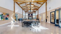 This single-story community amenity center is defined by its dramatic architectural centerpiece: a soaring vaulted ceiling supported by exposed heavy timber beams, illuminated by a series of globe chandeliers that cast warm, ambient light throughout the expansive main hall. Polished concrete floors reflect abundant natural light streaming in through rows of steel-framed windows positioned at both ground and clerestory levels, creating an airy, open atmosphere.

The great room serves as the social hub, featuring a large-format fireplace flanked by a mounted display screen, a leather seating lounge, and generous dining and gathering tables. Adjacent to this space, a sleek bar and kitchen island finished in deep navy cabinetry provides a functional entertaining area.

A dedicated mail and lounge room offers herringbone wood floors, teal-painted walls, vibrant yellow mailboxes, and a stylish chandelier overhead. A compact private office nook includes built-in wall-mounted paper roll workstations and geometric-patterned flooring, complemented by a separate conference-style room with dark built-in shelving and a display screen.

The fully equipped fitness center features wood-look flooring, teal accent walls, ceiling fans, and expansive windows, housing a comprehensive selection of cardio and strength training equipment. Together, these spaces form a cohesive, well-appointed amenity facility designed for community living.