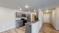 This single-story, 2-bedroom, 2-bathroom home is designed for comfortable, everyday living with a cohesive modern aesthetic carried throughout. The open-concept kitchen and living area serves as the heart of the home, featuring dark, rich cabinetry with a complementary granite-style countertop, black appliances including a range and built-in microwave, and a peninsula with a sink that doubles as a breakfast bar. Recessed lighting illuminates the kitchen, while wood-look luxury vinyl plank flooring flows seamlessly into the living area, which is brightened by a large sliding glass door fitted with vertical blinds. Ceiling fans are installed in the living and bedroom areas, adding both function and comfort. A wide hallway connects the main living spaces to the private quarters, where two bedrooms offer clean, neutral walls and generous closet storage with wire shelving systems. The primary bedroom is spacious and well-proportioned. Both bathrooms mirror the kitchen's design with matching cabinetry and granite-style countertops; one features a tub-shower combination with a curved rod and matte black fixtures. A dedicated laundry room includes utility hookups and overhead wire shelving for additional storage, rounding out this practical and well-finished home.