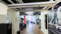 This fitness center is a purpose-built workout facility designed to accommodate a range of exercise needs. The space features an industrial-style ceiling with exposed ductwork, pipes, and flush panel lighting that illuminate the hardwood-style flooring throughout. The open layout provides ample room to move between equipment stations without feeling cramped.

The cardio zone is well-stocked with machines including stair climbers and treadmills, while a stability ball and balance accessories cater to functional training. A full-length wall mirror on one side enhances the sense of depth in the room while allowing users to monitor their form during workouts. Natural light filters in through windows along the far wall, contributing to a bright and energizing atmosphere.

Additional amenities include a wall-mounted magazine or media rack near the entrance, ideal for those who enjoy reading during cardio sessions. A wall clock keeps users on track with their routines. The space maintains a clean, utilitarian aesthetic with white walls and dark ceiling elements that give it a practical, no-frills character suited for focused training. Overall, this fitness center offers a compact yet comprehensive environment equipped for both cardiovascular and functional exercise.