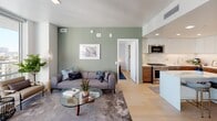 Located in the heart of downtown Los Angeles, this contemporary residence offers a thoughtfully designed open-concept layout that seamlessly connects the living, dining, and kitchen areas. Light hardwood floors run throughout, complementing the clean, modern aesthetic.

The kitchen is a standout feature, equipped with stainless steel appliances including a gas range, dishwasher, over-the-range microwave, and a large French door refrigerator. Two-tone cabinetry — white high-gloss uppers paired with warm wood lower cabinets — frames a white subway tile backsplash and bright countertops. A marble-topped kitchen island with bar seating adds both functionality and style.

The living area is anchored by sage green accent walls and large windows that bring in abundant natural light. An in-unit stacked washer and dryer is conveniently tucked away in a closet off the main living space.

The primary bedroom features floor-to-ceiling windows with direct access to a private outdoor balcony offering sweeping city views. A second bedroom also benefits from generous natural light. The bathroom is finished with large-format light tiles, a soaking tub with glass partition, a floating vanity, and open wood shelving, creating a clean, spa-like atmosphere.