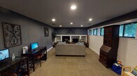 This single-floor space is thoughtfully designed to serve multiple functions, combining comfortable lounge areas with practical workspace zones. The interior is unified by deep navy-blue walls, dark ceilings, and recessed lighting throughout, creating a cohesive and polished ambiance.

The lounge area serves as the focal point of the space, anchored by a raised, tile-surround fireplace flanked by built-in shelving niches. A television is integrated into the hearth setup, and decorative ironwork wall art above adds a stylish touch. A plush sectional sofa faces the fireplace, complemented by a classic wooden coffee table. Neutral carpet tile flooring runs throughout, providing a warm, comfortable underfoot feel.

Along one wall, a row of low-profile windows allows natural light to filter in while offering views of outdoor greenery. A rich, dark-wood secretary bookcase stands nearby, adding traditional character. A dedicated computer workstation area features two desk setups against the blue accent wall.

A glass-and-steel partition wall separates a professional office corridor, visible through floor-to-ceiling glazing that maintains an open, connected feel. Near the entry, a chalkboard welcome sign and a hopscotch floor mat introduce a welcoming, playful element, rounding out this multifunctional space.