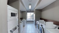 This single-floor laundry facility is designed for functionality and efficiency, offering a clean and organized environment for residents or guests. The room is lined on both sides with a combination of stacked and front-loading washing machines and dryers, providing ample capacity for multiple users simultaneously. The machines are arranged along the walls, leaving a clear central corridor for easy navigation and access.

The space features light gray walls accented with dark wood trim details near the countertop areas, lending a polished, modern aesthetic. Underfoot, large white ceramic floor tiles contribute to a bright and clean atmosphere. Overhead, recessed fluorescent lighting ensures the room is well-lit throughout, complemented by a central window at the far end of the room that allows natural light to filter in and offers a pleasant view of the outdoors.

The ceiling features subtle tray-style detailing with crown molding, elevating the overall design beyond that of a standard utility room. The layout is thoughtfully organized, maximizing the number of machines while maintaining an open, accessible feel. Overall, this laundry room balances practicality with a clean, well-maintained aesthetic.