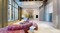 Located at 345 St Peter St in downtown Saint Paul, Minnesota, this sophisticated commercial lobby offers a refined and welcoming first impression. The single-floor space is generously proportioned, with soaring ceilings that amplify the sense of openness and grandeur. Floor-to-ceiling glass curtain walls flood the interior with abundant natural light while framing views of the surrounding urban streetscape, and a polished brass revolving entry door serves as a striking architectural focal point at the main entrance.

The flooring transitions elegantly between light travertine-toned stone in the main lobby and richly veined dark marble in the elevator corridor, delineating distinct zones within the space. A sculptural reception desk finished in dramatic blue-grey marble commands attention near the elevator bank, complemented by warm ambient underlighting. Overhead, contemporary ring-shaped pendant light fixtures add a modern artistic element against the crisp white ceiling.

A curved, jewel-toned sectional sofa in deep rose and burgundy anchors the seating lounge, paired with minimalist side tables in black and marble. Toward the rear, an ornate elevator lobby corridor features arched architectural detailing, decorative greenery, and refined wall treatments. A digital display panel is mounted flush to a warm wood-toned wall, adding a functional modern touch. The overall ambiance is polished, contemporary, and professional.