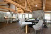 This impressive single-floor space is defined by its striking architectural design, most notably the soaring vaulted ceilings adorned with exposed natural wood timber beams and trusses that run throughout the main areas, creating a warm yet grand atmosphere. Rich hardwood-style flooring flows seamlessly across the entire space, complementing the bright white walls and abundant natural light that pours in through large windows and glass doors.

The central lounge and gathering area serves as the heart of the space, anchored by a bold, floor-to-ceiling geometric patterned tile fireplace wall that adds a sophisticated focal point. This area is thoughtfully furnished with tufted accent chairs, soft seating, and elegant marble-topped tables with gold bases, offering a comfortable and stylish setting for relaxation or informal meetings.

The open-plan work area features multiple workstations and is flanked by a glass-walled private conference room, providing both collaborative and focused work environments. Tall windows throughout ensure the workspace remains light-filled and connected to the lush greenery visible outside.

A sleek, modern kitchenette is also part of the space, outfitted with stone countertops, gray cabinetry, subway tile backsplash, and full-size stainless steel appliances. A corridor leads to additional rooms, completing this versatile and thoughtfully designed space.