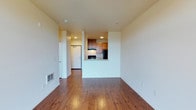 This single-floor unit offers a thoughtfully designed open layout that maximizes the sense of space and functionality. The living area is anchored by warm, rich hardwood flooring that extends throughout, creating a cohesive and inviting atmosphere. Clean white walls and recessed lighting contribute to a bright, well-lit environment that feels open and airy.

The kitchen is positioned at the heart of the unit, defined by a pass-through cutout that connects it seamlessly to the living space. Medium-toned wood cabinetry lines the kitchen walls, complemented by black appliances including a refrigerator and range. The pass-through design enhances the open feel of the space while providing a natural division between cooking and living areas.

A hallway leads from the main living space toward additional rooms, with a white entry door visible at the end, providing a clear sense of flow and circulation throughout the unit. A floor vent is integrated along the baseboard, reflecting practical utility details built into the design. The overall aesthetic is clean, modern, and functional, with neutral tones and natural wood finishes working together to create a comfortable and versatile living environment.