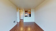 This single-floor unit offers a thoughtfully designed open layout that maximizes the sense of space and functionality. The living area is anchored by warm, rich hardwood flooring that extends throughout, creating a cohesive and inviting atmosphere. Clean white walls and recessed lighting contribute to a bright, well-lit environment that feels open and airy.

The kitchen is positioned at the heart of the unit, defined by a pass-through cutout that connects it seamlessly to the living space. Medium-toned wood cabinetry lines the kitchen walls, complemented by black appliances including a refrigerator and range. The pass-through design enhances the open feel of the space while providing a natural division between cooking and living areas.

A hallway leads from the main living space toward additional rooms, with a white entry door visible at the end, providing a clear sense of flow and circulation throughout the unit. A floor vent is integrated along the baseboard, reflecting practical utility details built into the design. The overall aesthetic is clean, modern, and functional, with neutral tones and natural wood finishes working together to create a comfortable and versatile living environment.
