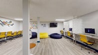 Located at 914 W Lake St in Fort Collins, Colorado, this dynamic single-floor workspace is thoughtfully designed to support a variety of working and collaborative styles. The open layout features light wood-grain flooring, white walls, and flush linear lighting throughout, creating a bright and welcoming atmosphere.

The main area serves as a flexible common space, outfitted with oversized bean bag chairs in navy and yellow, providing casual seating options alongside more structured workstations. A wall-mounted flat-screen TV and colorful wall art contribute to the space's lively, energetic character. A workstation zone lines one wall, featuring a row of iMac computers set along a sleek desk surface, separated from the open area by striking white geometric sculptural panels that double as stylish dividers.

A long communal table with rolling yellow chairs offers a dedicated area for group work or presentations, accented by a geometric diamond wall art piece above. A round white pedestal table with blue velvet chairs anchors a meeting nook, set atop a vibrant teal, yellow, and grey area rug.

The kitchenette is finished with white shaker cabinetry, stone-look countertops, a stainless steel sink, microwave, and a bold teal glass tile backsplash that adds a pop of color. A full-size stainless steel refrigerator completes the space. Together, these elements make this a well-equipped, design-forward environment ideal for collaboration and focused work.