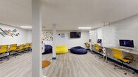 Located at 914 W Lake St in Fort Collins, Colorado, this dynamic single-floor workspace is thoughtfully designed to support a variety of working and collaborative styles. The open layout features light wood-grain flooring, white walls, and flush linear lighting throughout, creating a bright and welcoming atmosphere.

The main area serves as a flexible common space, outfitted with oversized bean bag chairs in navy and yellow, providing casual seating options alongside more structured workstations. A wall-mounted flat-screen TV and colorful wall art contribute to the space's lively, energetic character. A workstation zone lines one wall, featuring a row of iMac computers set along a sleek desk surface, separated from the open area by striking white geometric sculptural panels that double as stylish dividers.

A long communal table with rolling yellow chairs offers a dedicated area for group work or presentations, accented by a geometric diamond wall art piece above. A round white pedestal table with blue velvet chairs anchors a meeting nook, set atop a vibrant teal, yellow, and grey area rug.

The kitchenette is finished with white shaker cabinetry, stone-look countertops, a stainless steel sink, microwave, and a bold teal glass tile backsplash that adds a pop of color. A full-size stainless steel refrigerator completes the space. Together, these elements make this a well-equipped, design-forward environment ideal for collaboration and focused work.