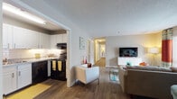This single-floor residence offers a thoughtfully designed layout that balances comfort and functionality. The open-concept living area serves as the heart of the home, featuring warm hardwood-style flooring, large windows that invite natural light, and a spacious feel enhanced by a textured ceiling. The living room is anchored by a media console and a mid-century-inspired coffee table, complemented by vibrant accent colors from bold curtains and decorative rugs that add personality to the space.

Adjacent to the living area, the kitchen is efficiently laid out with crisp white cabinetry, granite-style countertops, a classic subway tile backsplash, and black appliances including a range and dishwasher. Under-cabinet lighting adds a functional and welcoming ambiance.

A hallway connects the main living spaces to two well-proportioned bedrooms, each featuring plush carpeting, windows with outdoor views, and ample wall space. The bedrooms are styled with colorful artwork and warm-toned furnishings, creating inviting retreats.

The full bathroom is finished with wood-look tile flooring, a white vanity with stone-style countertops, and a shower-tub combination with a curtain enclosure. Neutral wall tones throughout the unit create a cohesive backdrop that ties each room together seamlessly.
