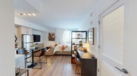 Located at 112 NE Third St in Pompano Beach, Florida, this thoughtfully designed studio unit makes efficient use of its single-floor layout. The open-plan main area seamlessly integrates the living, sleeping, and dining zones, unified by warm-toned hardwood-style flooring and crisp white walls that enhance the sense of brightness throughout. A freestanding open shelving unit serves as a functional room divider, subtly separating the sleeping area from a dedicated workspace with a built-in desk. The sleeping area is anchored by a full-size bed positioned beneath a large window dressed with sheer curtains and blinds, allowing generous natural light to fill the space while offering privacy. The kitchen is well-appointed with light wood cabinetry, open shelving, a subway tile backsplash, and a full suite of stainless steel appliances including a refrigerator, range, microwave, and dishwasher. A small dining table provides a functional eating area just steps from the kitchen. The bathroom features tile flooring, a full-size soaking tub with a curved shower rod, and clean white finishes throughout. A separate closet with louvered doors adds practical storage. The unit's clean, modern aesthetic and efficient design make it a functional and inviting living space.