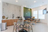 Located at 199 Peters St SW in Atlanta, Georgia, this single-floor space is designed as a versatile communal gathering area with a clean, contemporary aesthetic. The layout is thoughtfully divided into functional zones that flow seamlessly into one another across light-toned tile flooring throughout.

The lounge area anchors one end of the space, featuring a deep navy upholstered banquette set against a bold forest-green accent wall. Industrial-style pendant bulb lighting hangs above, adding warmth and character. Accompanying armchairs and round wooden side tables create an inviting atmosphere suited for casual conversation or relaxed gatherings. Natural light filters in through multiple windows fitted with horizontal blinds, offering views of the surrounding urban exterior.

Adjacent to the lounge, a dining zone centers around a round marble-topped table surrounded by patterned upholstered chairs. A wall-mounted flat-screen television provides additional functionality for presentations or entertainment. The kitchenette along one wall features warm wood cabinetry, a sleek countertop, and a black matte sink fixture, backed by an artistically textured accent wall that adds visual interest.

A separate anteroom near the entrance provides seating alongside restroom access via a clearly marked door, and a glass-paneled door connects to an adjacent space, enhancing the overall openness of the layout.