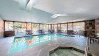 This single-level indoor aquatic facility is thoughtfully designed to offer year-round recreational use in a bright, welcoming environment. The centerpiece is a large rectangular swimming pool with clear turquoise water, finished with tiled edges and accessed via built-in entry steps with stainless steel handrails. Adjacent to the pool, an octagonal hot tub is recessed into the slate-toned tile floor, fitted with jets and its own entry rail, providing a dedicated relaxation area.

The architecture features soaring vaulted ceilings with multiple skylights that flood the space with abundant natural light, enhancing the openness of the interior. Louvered wood-panel windows line the perimeter walls, allowing for natural ventilation and views of the surrounding greenery while maintaining a sense of enclosure. Stone veneer accent walls add a warm, lodge-like aesthetic throughout the facility.

A spacious pool deck surrounds the water on all sides, furnished with lounge chairs and side tables arranged to encourage relaxation and socialization. A dedicated seating area near a translucent glass-block wall houses additional chairs and provides separation from the pool area. The facility also includes a utility and entry area with safety equipment and signage, ensuring both functionality and comfort throughout the space.