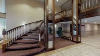 Located at 25400 Rockside Rd in Bedford, Ohio, this well-appointed single-floor commercial space makes a strong first impression with its stately and traditional interior design. The centerpiece of the space is a graceful curved staircase adorned with rich dark wood banisters and white spindle balusters, accented by plush patterned carpet in deep burgundy tones. Full-length mirrors flanking the staircase create a sense of expanded space and reflect the elegant architectural details, including a chandelier that adds a refined, formal touch overhead. The flooring transitions from warm beige tile in the entry area to carpeted surfaces in the surrounding spaces, offering both practicality and comfort underfoot. Neutral, softly lit walls in warm taupe complement the dark wood millwork and contribute to a calm, dignified ambiance throughout. Decorative greenery and a cushioned bench provide welcoming accents that enhance the approachable character of the interior. The drop ceiling with recessed lighting ensures consistent, even illumination across the space. The overall atmosphere is composed and professional, evoking a sense of quiet formality well-suited for a reception or gathering-oriented use. The thoughtful blend of classic architectural elements and understated décor makes this a distinguished and functional space.