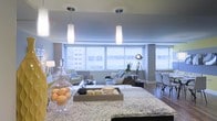 This single-floor space showcases a thoughtfully designed open-concept layout that effortlessly connects the kitchen, dining, and living areas into one cohesive and functional environment. The kitchen features a generous granite-topped island with striking veined stone detailing, providing ample prep space and a natural focal point for the room. Elegant pendant lights with frosted glass shades hang above, casting a warm, inviting glow that complements the overall ambiance. The living area is anchored by a comfortable sectional sofa and accented with a bold industrial-style tripod floor lamp, adding a modern edge to the space. Large windows with horizontal blinds span the width of the room, allowing abundant natural light to flood the interior while offering expansive views of the surrounding cityscape. The dining area sits adjacent to the living space, furnished with sleek contemporary chairs that maintain the modern aesthetic. Warm hardwood flooring flows throughout, tying the open layout together with a sense of warmth and continuity. Cool grey-blue wall tones complement the neutral furnishings, creating a calm, sophisticated atmosphere well-suited for both everyday living and entertaining.