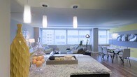 This single-floor space showcases a thoughtfully designed open-concept layout that effortlessly connects the kitchen, dining, and living areas into one cohesive and functional environment. The kitchen features a generous granite-topped island with striking veined stone detailing, providing ample prep space and a natural focal point for the room. Elegant pendant lights with frosted glass shades hang above, casting a warm, inviting glow that complements the overall ambiance. The living area is anchored by a comfortable sectional sofa and accented with a bold industrial-style tripod floor lamp, adding a modern edge to the space. Large windows with horizontal blinds span the width of the room, allowing abundant natural light to flood the interior while offering expansive views of the surrounding cityscape. The dining area sits adjacent to the living space, furnished with sleek contemporary chairs that maintain the modern aesthetic. Warm hardwood flooring flows throughout, tying the open layout together with a sense of warmth and continuity. Cool grey-blue wall tones complement the neutral furnishings, creating a calm, sophisticated atmosphere well-suited for both everyday living and entertaining.