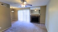 Located in Irving, Texas, this single-floor residence offers a comfortable and functional layout designed for everyday living. The living room serves as the heart of the home, immediately drawing attention with its classic brick fireplace topped by a decorative mirror and white mantel — a charming focal point that adds warmth and character to the space. A ceiling fan with integrated lighting is centrally positioned overhead, ensuring year-round comfort and adequate illumination. Large vertical blinds frame a full-width sliding glass door that invites abundant natural light into the room, creating a bright and airy atmosphere. Plush wall-to-wall carpeting in a neutral tone runs throughout the living area, contributing to a soft and welcoming underfoot feel. The walls are finished in a warm beige tone, complementing the earthy brick of the fireplace and tying the room's aesthetic together cohesively. A textured ceiling adds a subtle traditional touch to the overall interior. The space is thoughtfully arranged to balance comfort and practicality, making it well-suited for both relaxing and entertaining. The single-floor plan offers ease of movement and accessibility throughout.