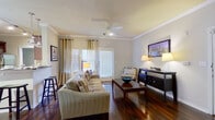 This single-floor residence offers a cohesive and thoughtfully designed living space that balances style and functionality throughout. The open-concept living room is anchored by rich, dark hardwood flooring, elegant crown molding, and a ceiling fan that add warmth and architectural character. Multiple windows dressed with layered curtains allow natural light to fill the room, while a set of doors provides access to the outdoors. A dedicated dining area adjacent to the living space features a window with decorative curtains, creating a bright and comfortable setting for meals. The kitchen is well-equipped with rich cherry wood cabinetry, granite countertops, a tiled backsplash, stainless steel appliances including a refrigerator, gas range, and microwave, plus a breakfast bar for casual dining. Track lighting illuminates the workspace effectively. The bedroom is a serene retreat, showcasing plush carpeting, crown molding, a ceiling fan, and generous natural light from a large window with green drape curtains. A spacious walk-in closet features built-in wire shelving and hanging rods for ample storage. The bathroom is finished with granite countertops, cherry wood cabinetry, a large vanity mirror, and a shower-tub combination.