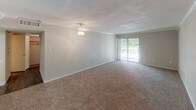 This single-floor residence offers a straightforward, functional layout across several well-defined spaces. The living area is generously sized, finished with wall-to-wall carpeting and neutral-toned walls that create a bright, airy atmosphere. Crown molding adds a refined touch, while a ceiling fan with a light fixture provides both comfort and illumination. A large sliding glass door floods the room with natural light and opens to an outdoor area, offering views of surrounding greenery. An open pass-through connects the living space to the kitchen, enhancing the sense of openness.

The galley-style kitchen is efficiently arranged, featuring white cabinetry on both sides, a double-basin stainless steel sink, and a white electric range. A built-in microwave is mounted above the stove, and the dark hardwood-style flooring contrasts cleanly with the white finishes. A dedicated pantry with built-in shelving provides ample storage.

The bathroom continues the dark hardwood-style flooring and maintains the clean, white aesthetic throughout. It includes a full soaking tub with a showerhead and chrome fixtures, a wall-mounted towel bar, and a toilet, all accented by recessed ceiling lighting.